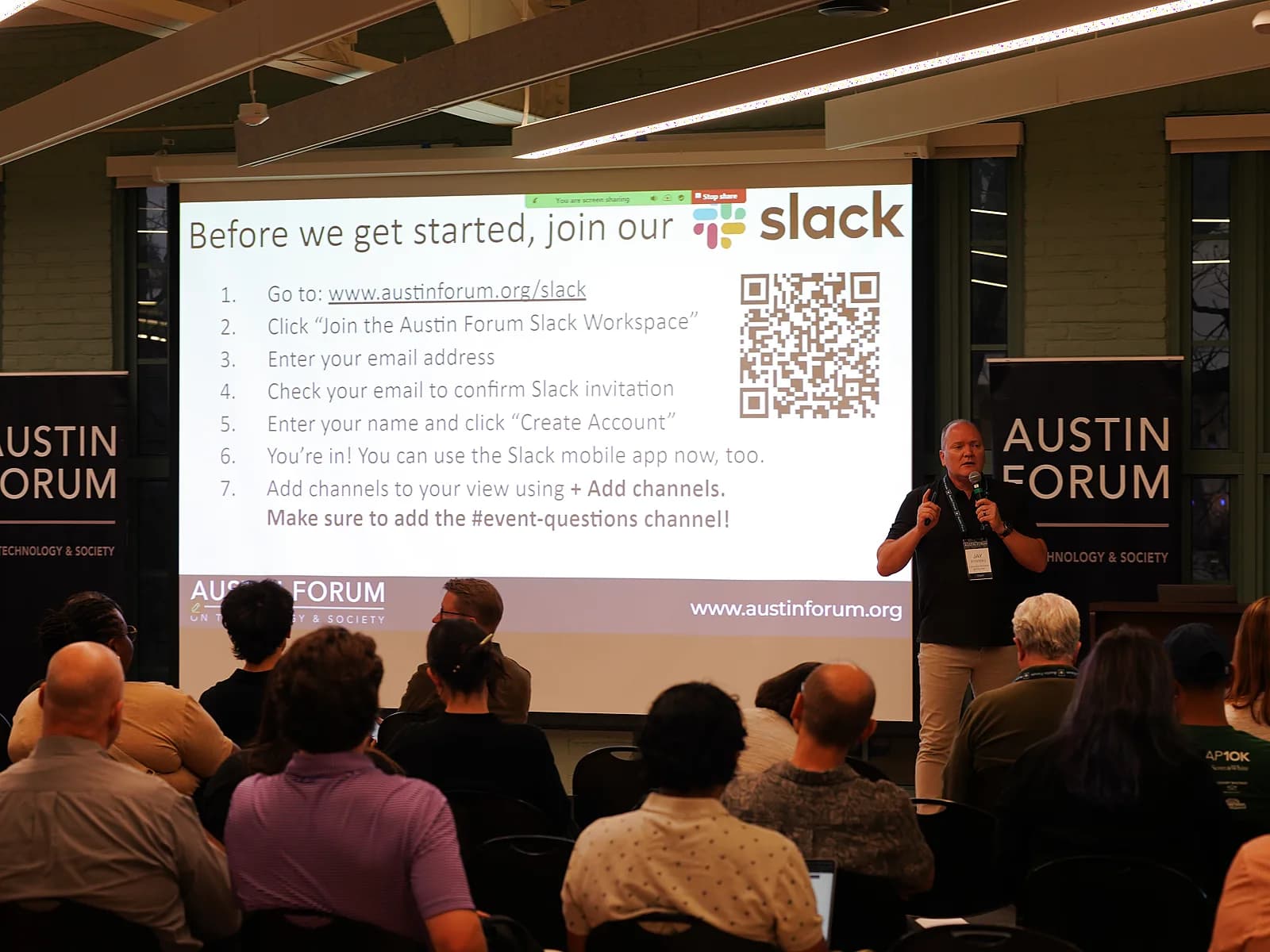 A full room of attendees seated facing a panel on stage beneath an Austin Forum banner, with a speaker at the lectern